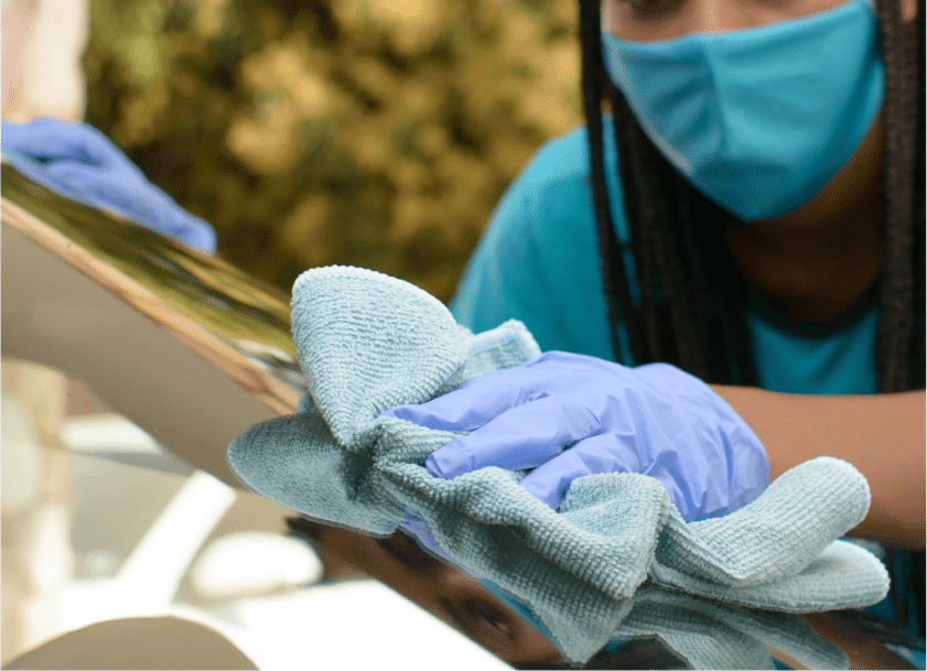 Pessoa usando blusa, máscara de proteção e luvas na cor azul. Utiliza um tecido próprio para limpar o para-brisa de um veículo. Imagem ilustrataiva do texto estética automotiva Maranhão.