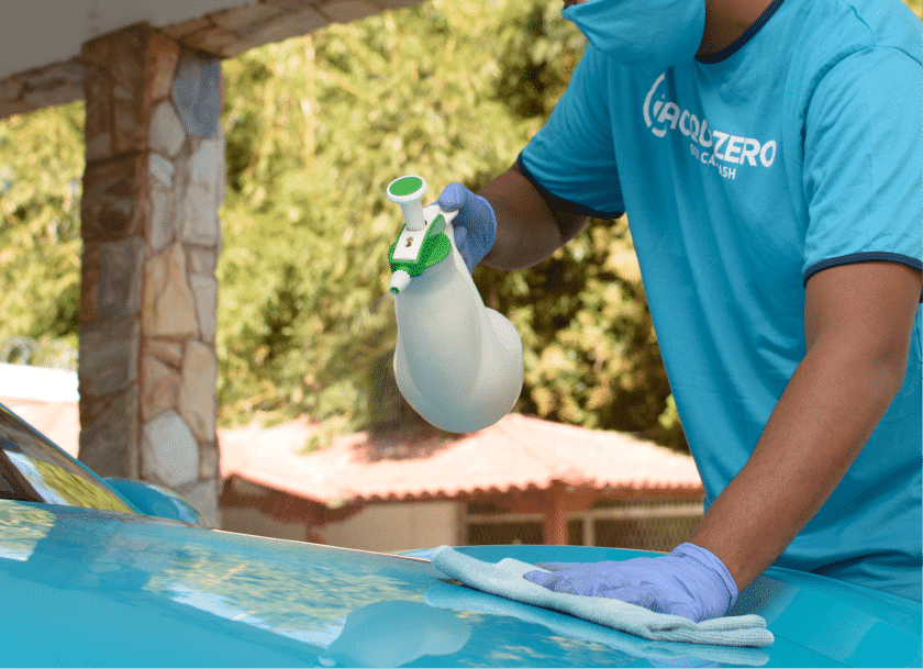 Funcionário da Acquazero usando blusa e luvas na cor azul claro, limpando o capô de um carro também azul com o auxílio de uma flanela e borrifador. Imagem ilustrativa do texto sobre lavagem a seco em Recife.