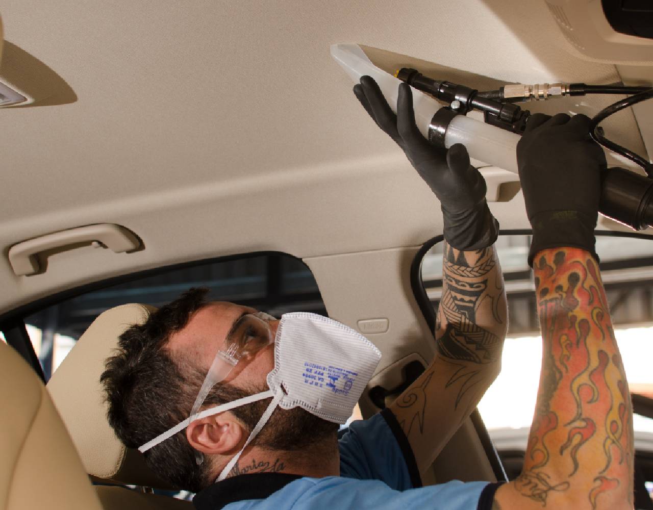 Homem trajando blusa azul e máscara de proteção branca realiza a limpeza do interior de um veículo com o auxílio de um dispositivo. Ilustração do texto lavar sofá Araraquara.