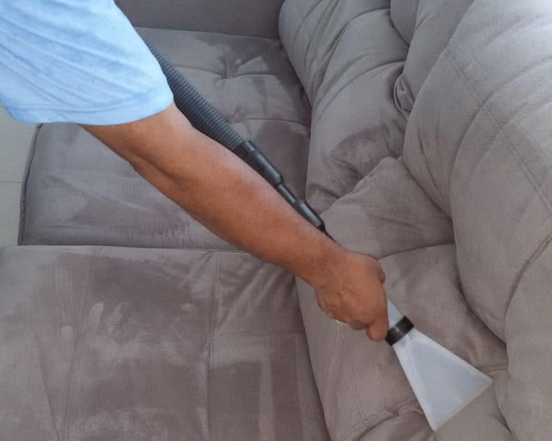 Pessoa higieniza um sofá cinza com o auxílio de um aspirador de pó. Ilustração do texto lavar sofá perto São Luís.