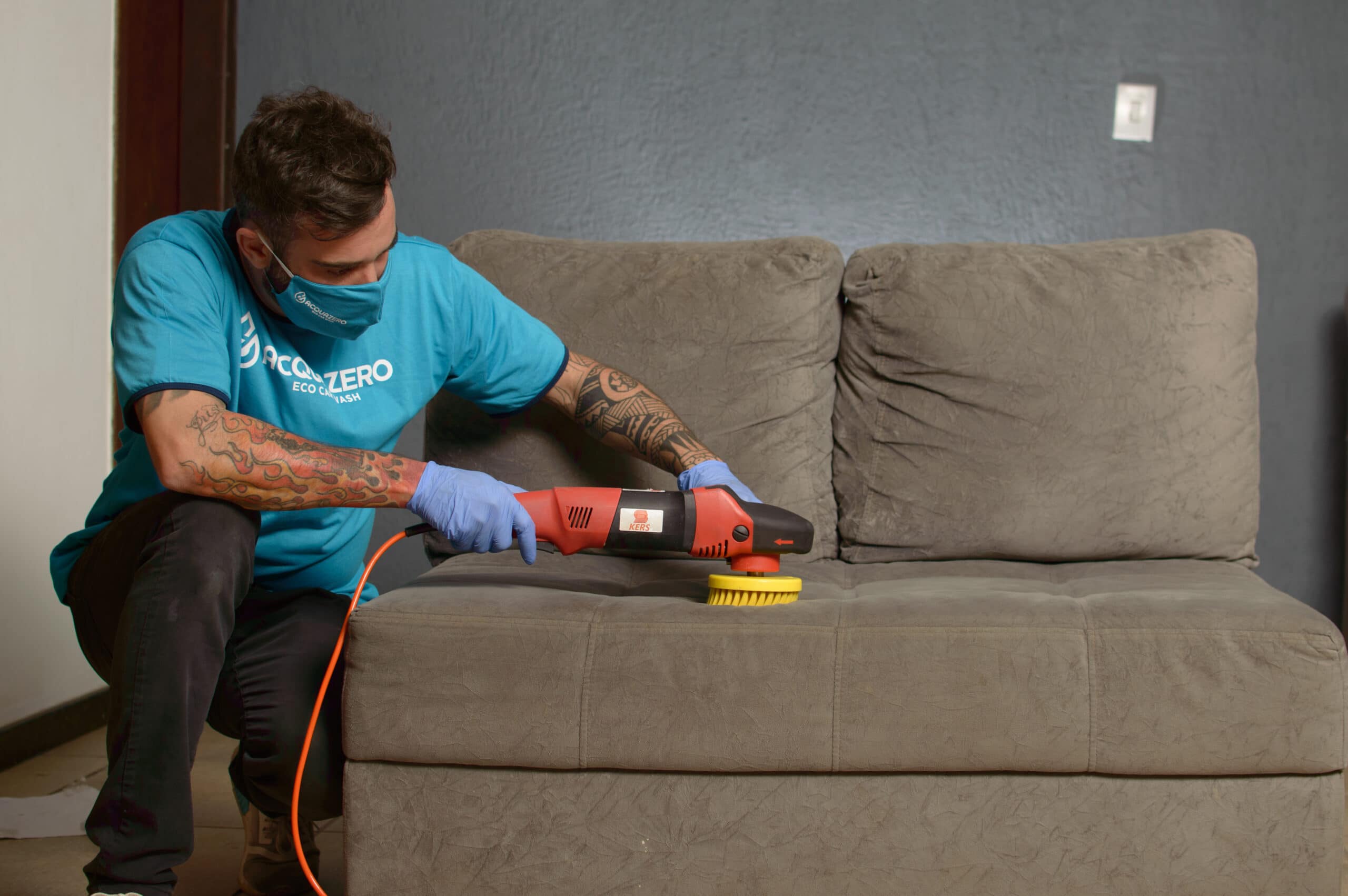 Técnico da Acquazero usando camisa e luvas azuis, higieniza o sofá com o apoio de uma escova elétrica amarela e laranja. Conteúdo do texto lavar sofá no Flamengo.
