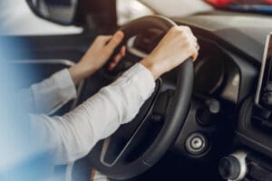 Imagem de braços de uma pessoa com camisa de manga longa, segurando o volante de um veículo.