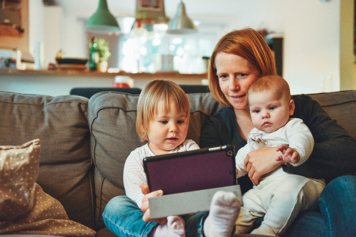 Mãe com criança e bebê, sentados no sofá com tablet. 