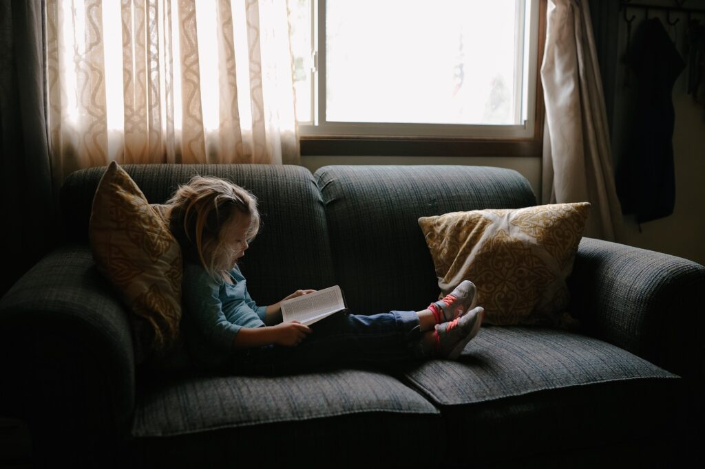 Uma menina criança de blusa azul, calça jeans e tênis, sentada em um sofá cinza, lendo um livro. Ao fundo uma janela com cortinas brancas. Imagem ilustrativa para o texto limpar sofá horto.