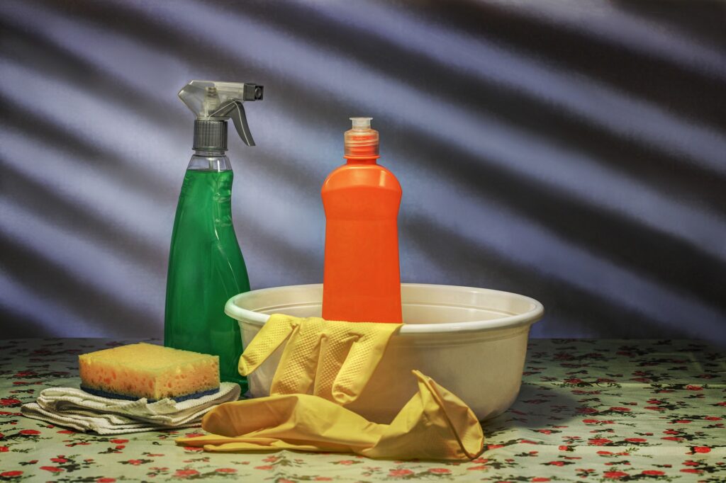 Imagem mostra uma bacia branca, com uma embalagem laranja dentro e luvas amarelas. Fora da bacia observa-se uma esponja e uma embalagem com um líquido verde. Ilustração do texto mistura para limpar colchão.