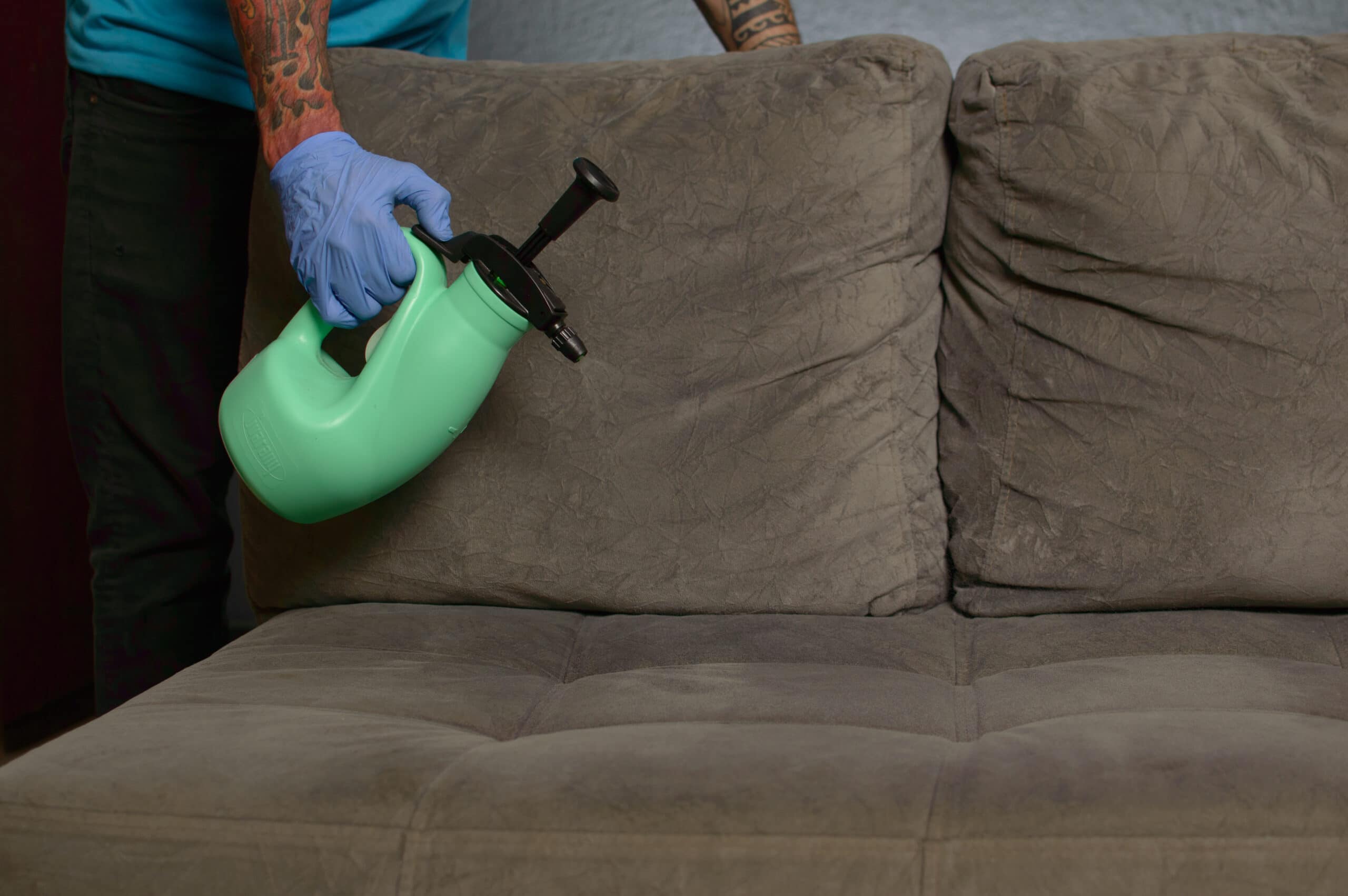 Homem com luva azul utiliza um borrifador verde para higienizar um sofá cinza. Ilustração do texto lavar sofá Centro de João Pessoa.