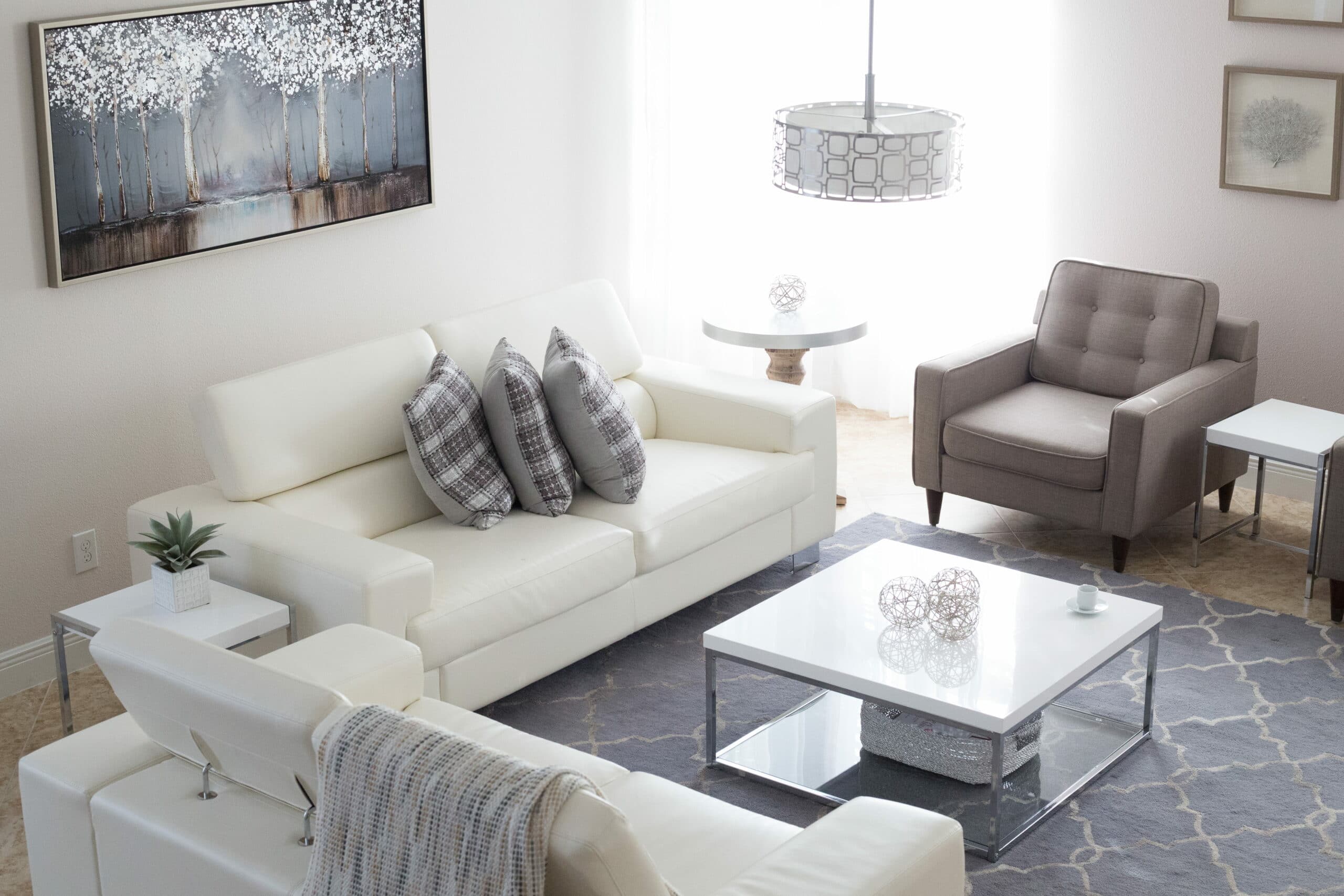 Quadro, lustre, sofás, amofadas, poltrona, tapete e mesa de centro, cinzas e brancos. Imagem do conteúdo sobre