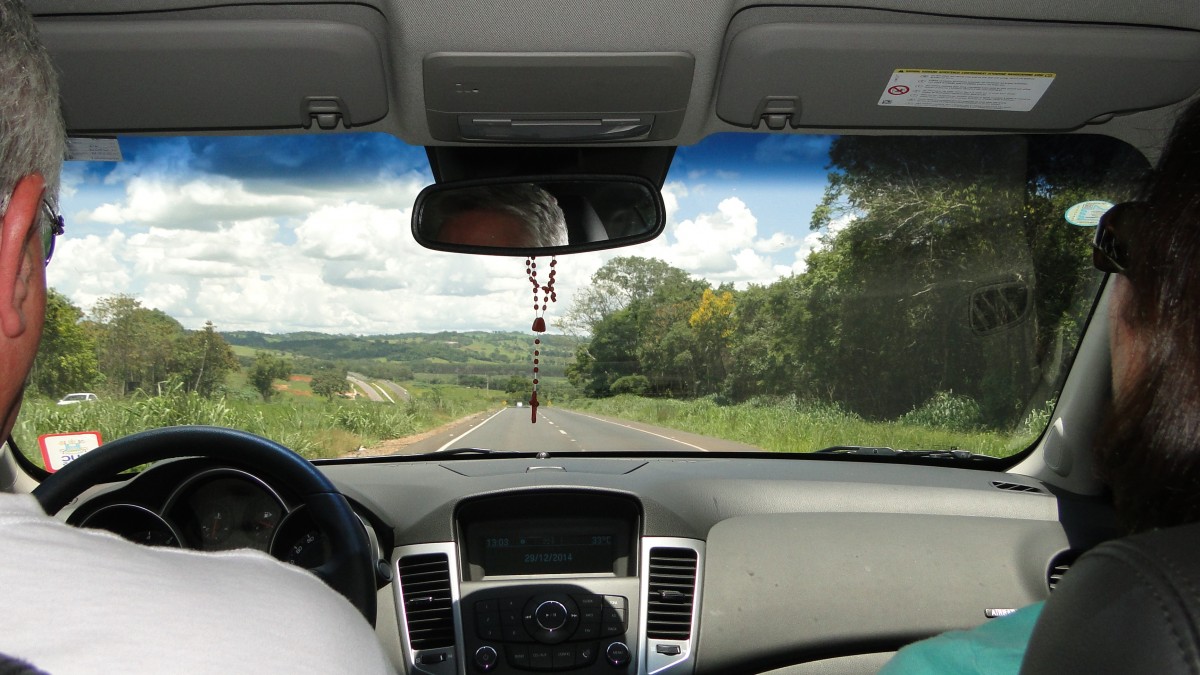 Visão de um carro por dentro, com motorista, terço no retrovisor e estrada. Imagem ilustrativa texto sanitização e desinfecção.