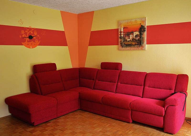 Sofá vermelho de canto, parede creme, vermalha e laranja, com relógio e quadro. Imagem do conteúdo lavar sofá Brotas.