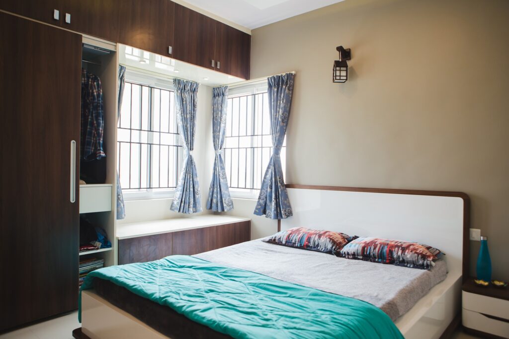 cama de casal em um quarto iluminado com uma grande janela e um guarda roupa. imagem de texto sobre como tirar mofo de roupa