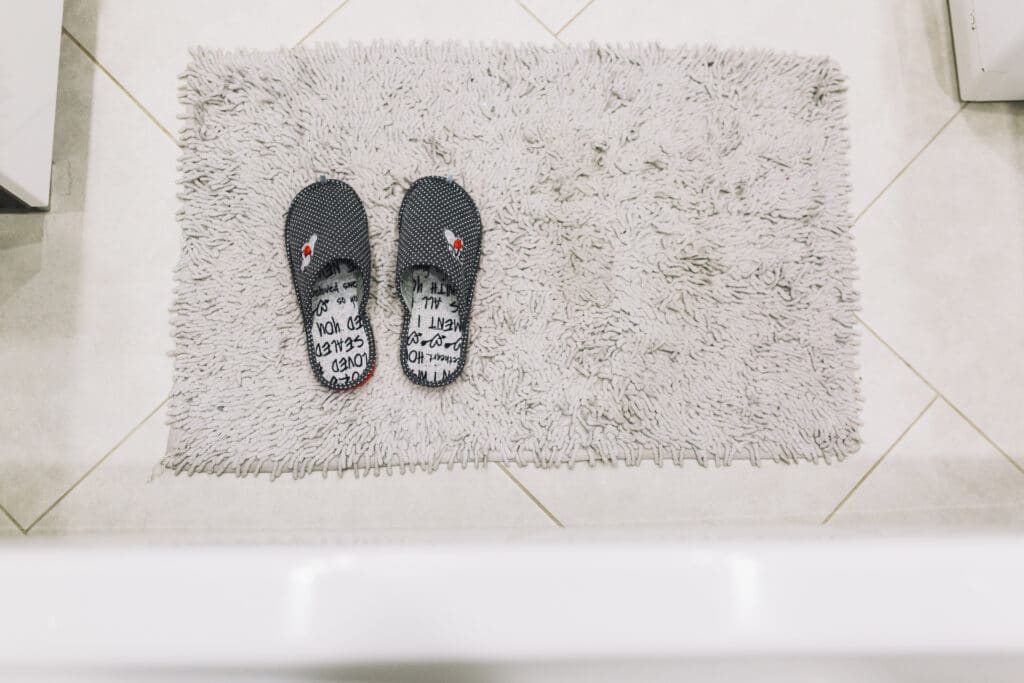 Dois chinelos em cima de um tapete felpudo branco no chão do banheiro. Imagem ilustrativa texto como lavar tapete felpudo.