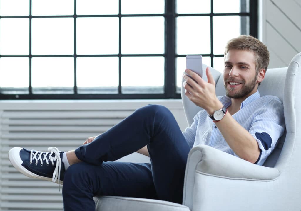 Homem sentado em uma poltrona branca e olhando o celular. Imagem ilustrativa texto lavar sofá no Promissão.