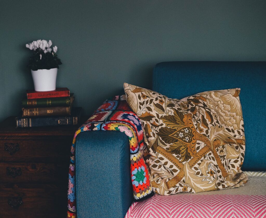 Braço de um sofá azul com uma almofada colorida, ao lado uma mesinha com livros empilhados. Imagem ilustrativa texto impermeabilização de sofá no Espírito Santo.