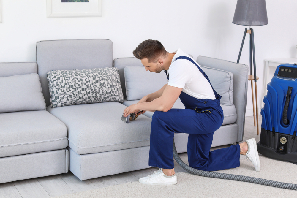 Homem fazendo a limpeza de um sofá grande e cinza com equipamento. Imagem ilustrativa do texto lavagem de sofá a seco.