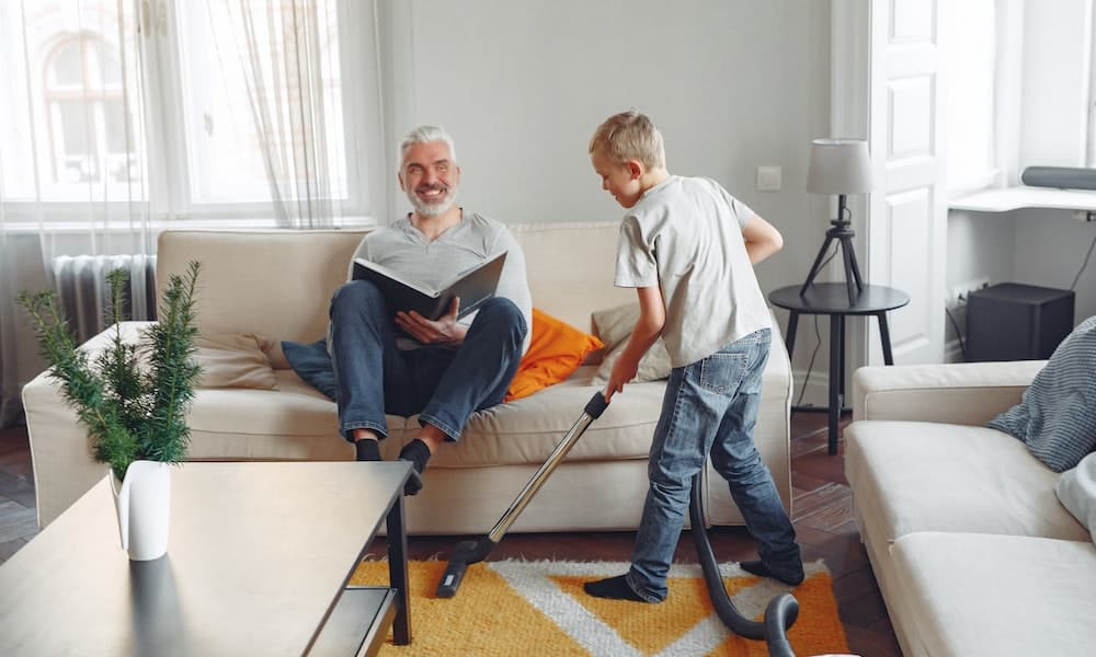 Homem de camisa cinza sentado no sofá levanta os pés para que uma criança com vassoura na mão possa limpar o chão. Imagem ilustrativa lavar sofa sao francisco.
