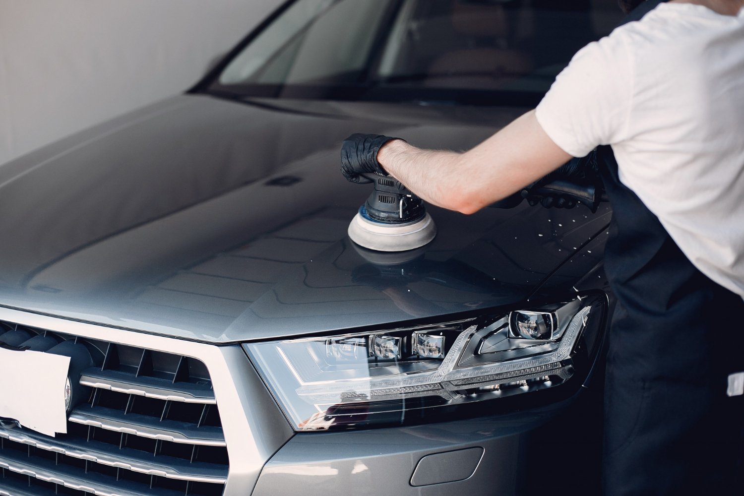 Pessoa usando blusa branco e avental preto, polindo um carro cinza com o auxílio de um dispositivo. Ilustração do texto sobre lavar o carro.