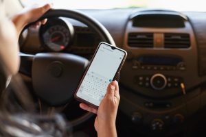 Mão esquerda uma mulher segura o volante enquanto mão direita digita em um celular. Imagem ilustrativa texto carro cheiroso