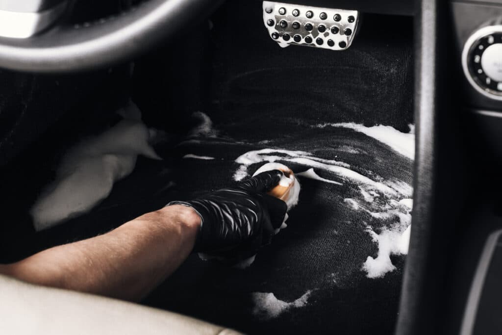Mão utilizando luva preta, higieniza o interior de um carro preto com o auxílio de uma escova. Há espuma no carpete do carro devido a limpeza realizada. Ilustração do texto como limpar o carpete do carro.