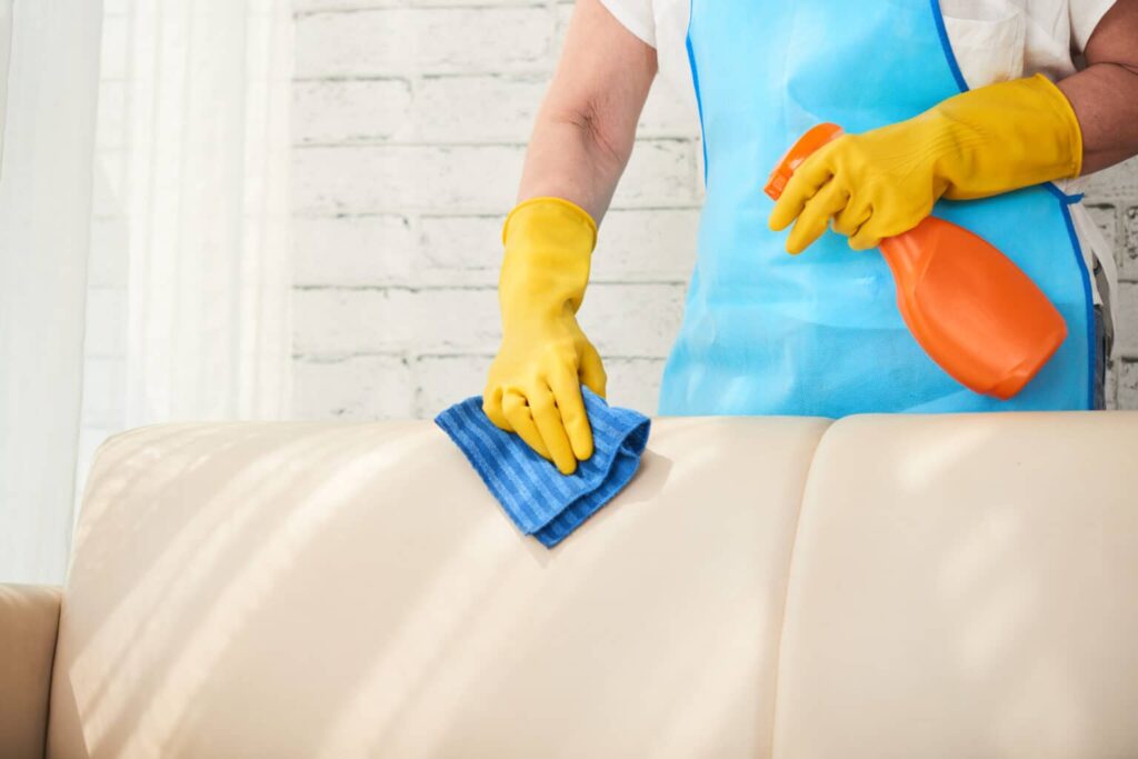 Image of a person with yellow glove and blue apron, cleaning a light sofa with the aid of an orange spray bottle and blue cloth.  Illustration of the text how to remove mold.