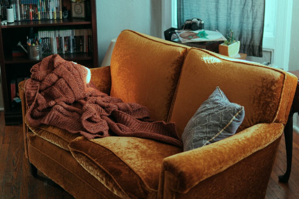 Sofá marrom de estofado com uma coberta em cima e uma almofada cinza. Imagem ilustrativa lavar sofá no Sagrada Família.