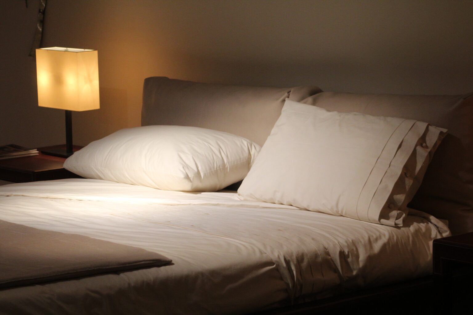 Colchão e travesseiro, com roupa de cama branca e bege. Abajur ao lado direito. Imagem ilustrativa do texto mancha de café do sofá.