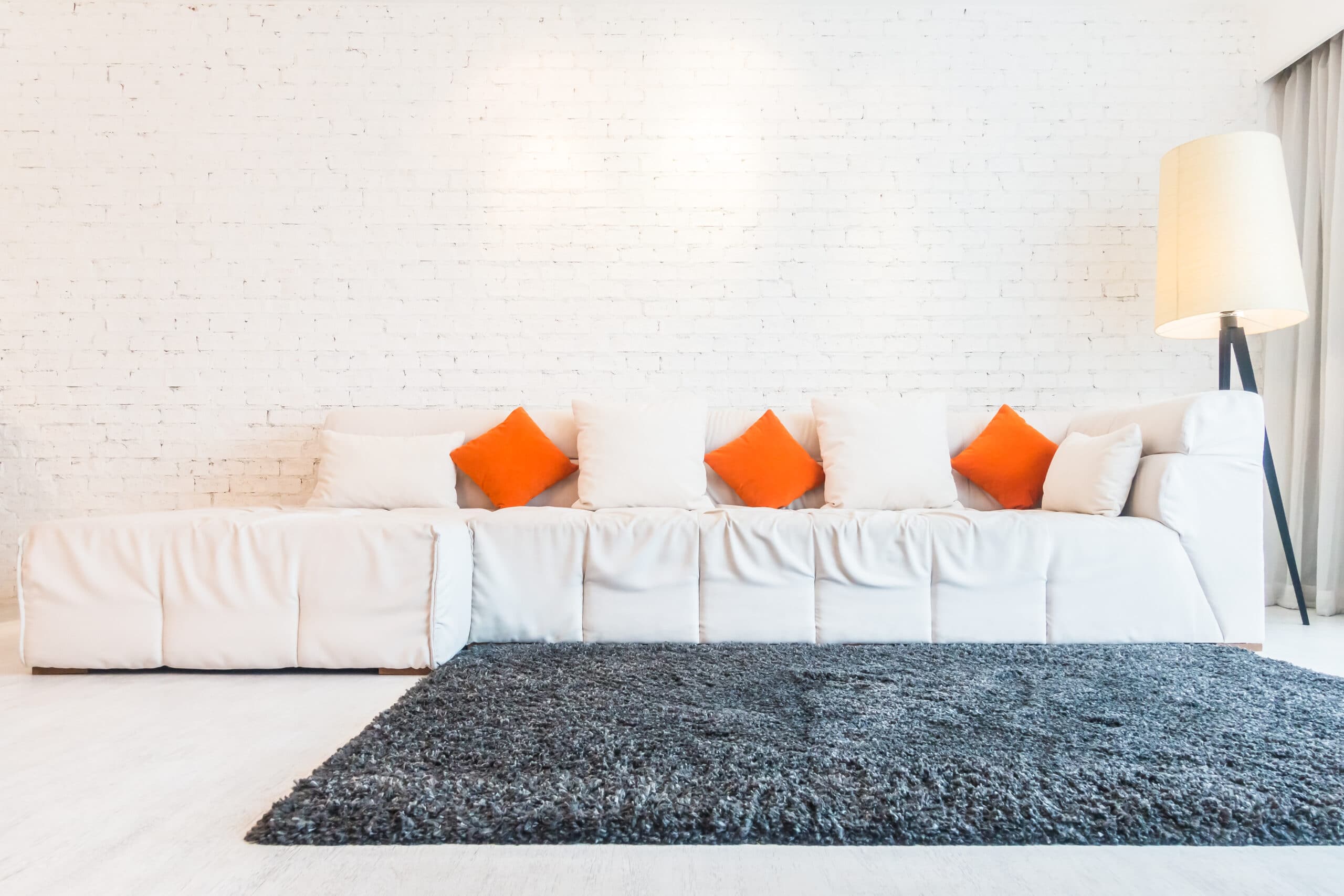 Imagem de uma sala de estar com tapete cinza, sofá claro e almofadas nas cores laranja e branco. Ilustração do texto como impermeabilizar tecido.