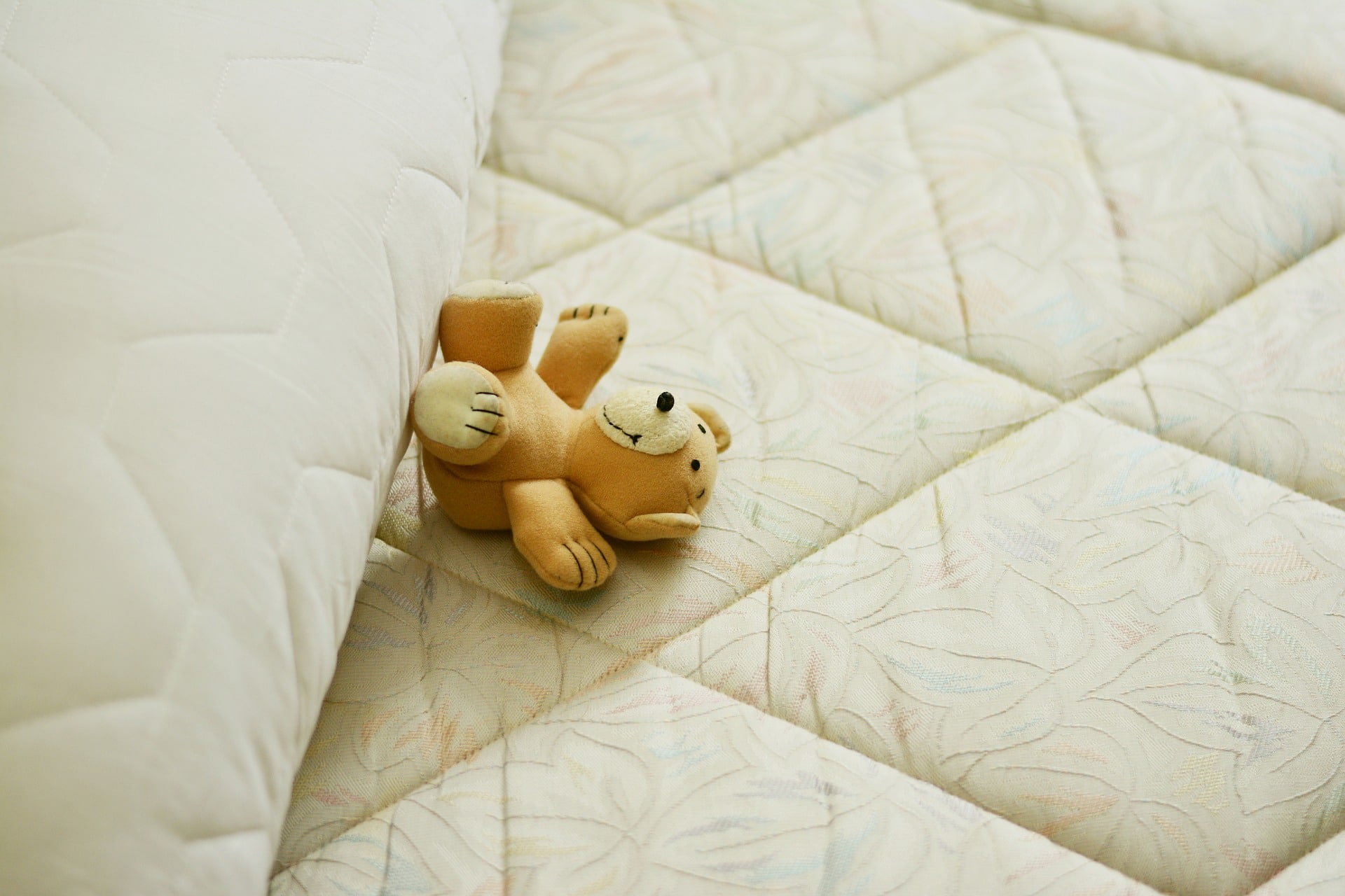 Imagem de um urso bege de ponta cabeça, sobre um colchão com tom claro. Ilustração do texto sobre como limpar colchão.