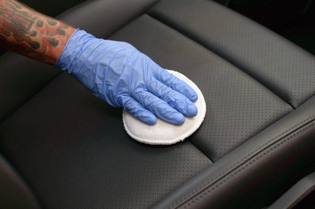 Mão com luva azul realiza a limpeza de um banco automotivo com o auxílio de um tecido branco. Ilustração do texto sobre como limpar couro sintético.