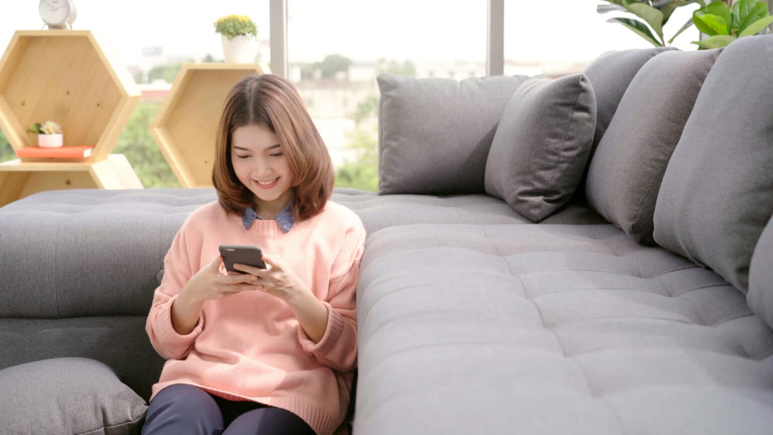 Mulher sentada ao lado de um sofá cinza mexendo no celular.