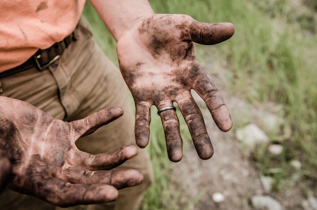 Hands dirty with grease, on a background with grass and a person wearing brown pants and orange blouse.  Illustrative image for the text how to remove grease stain.