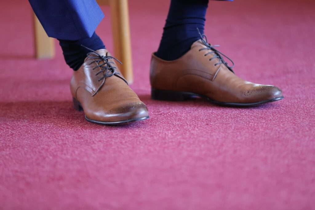 Pés masculinos com sapatos marrons em cima de um carpete vermelho. Ilustração do texto empresas de limpeza Caxias do sul.