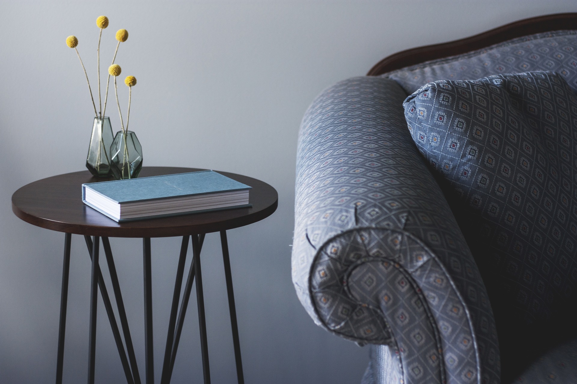 Braço de um sofá estampado e um móvel porta livro ao lado com um vaso de flor e um livro. Imagem do texto sobre limpar sofá no Centro de Contagem.