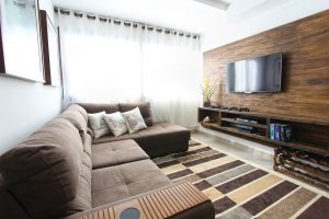 Sala com um sofá marrom em L com cinco lugares e uma televisão em um painel na parte à sua frente. Imagem ilustrativa texto como desinfetar superfície de tecido.