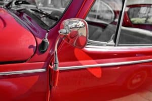 Imagem da lateral de um carro antigo na cor vermelha com um retrovisor cromado. Imagem ilustrativa texto aplicar cera no carro.