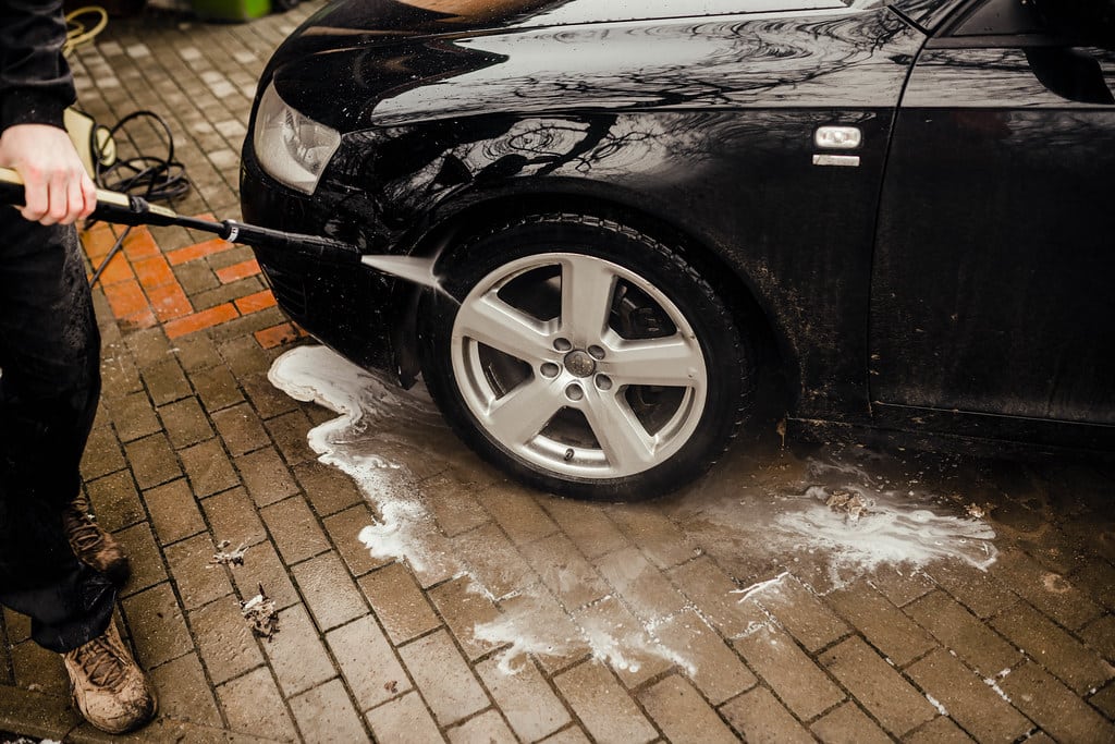 Homem lava carro preto com uma mangueira. Imagem ilustrativa texto dicas de limpeza automotiva.