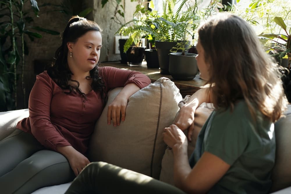 Duas mulheres conversando, sentadas em um sofá em cor bege claro. Ilustração do texto sobre como eliminar ácaros.