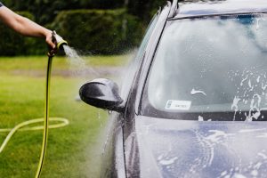 Imagem de uma mão segurando uma mangueira amarela jogando água em um carro escuro com espuma na lataria e nos vidros. Imagem ilustrativa texto como cuidar do carro em casa.