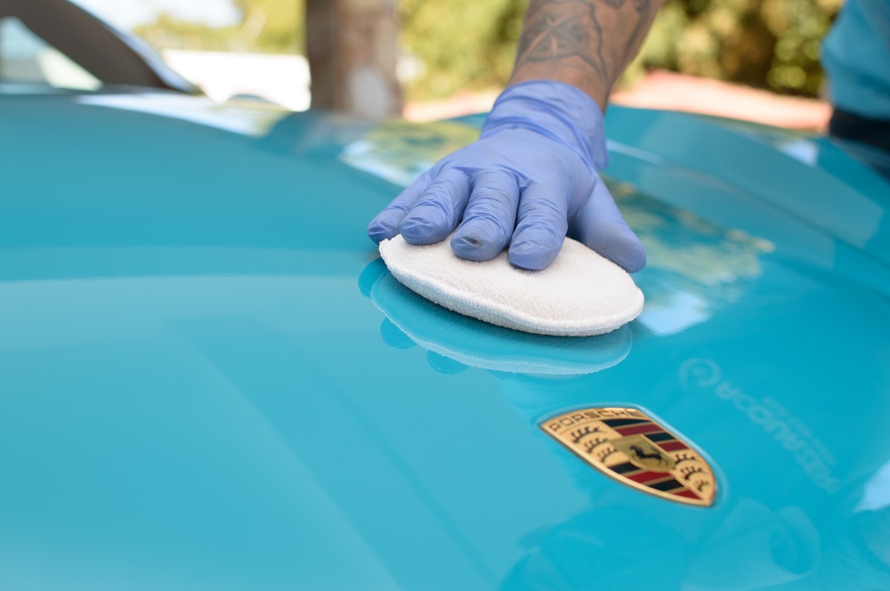 Mão usando luva azul limpando capo de um carro. Ilustração do texto sobre como fazer lavagem a seco automotiva.