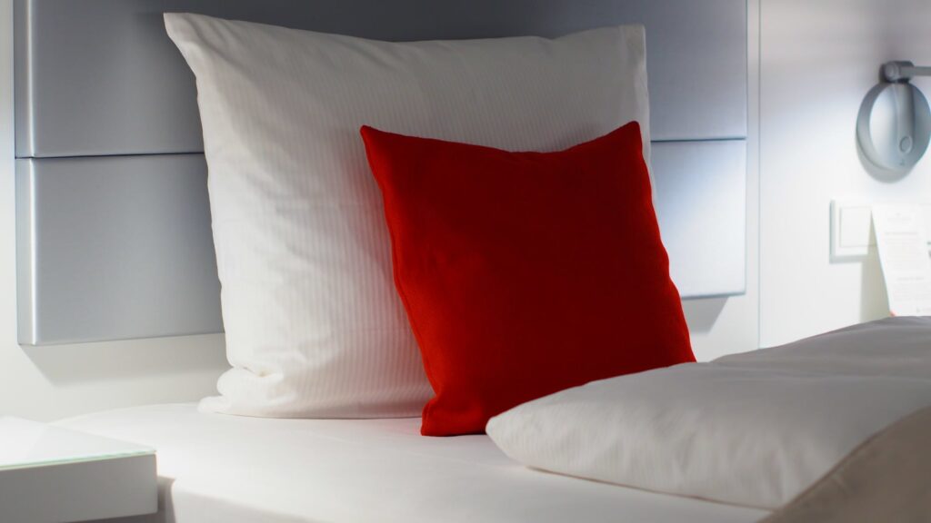 Dois travesseiros brancos e uma almofada vermelha na cama. Imagem ilustrativa do texto sobre como tirar manchas de travesseiros.