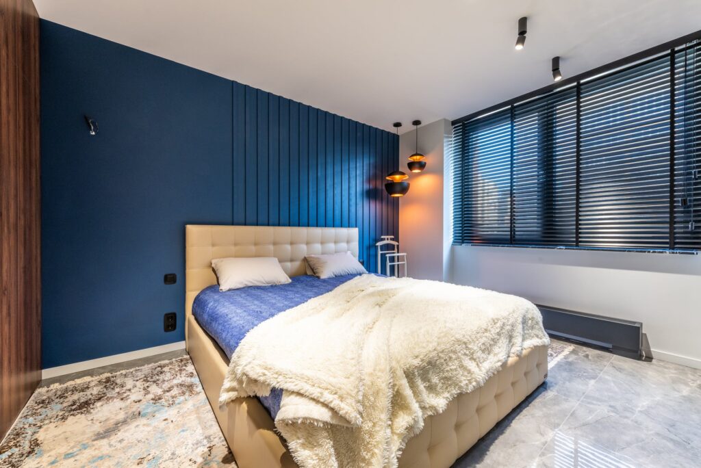 Quarto com cama de casal,, lençol azul sobre o colchão,cobertores felpudos bege e persiana fechada. Imagem ilustrativa do texto sobre limpar o colchão a seco.