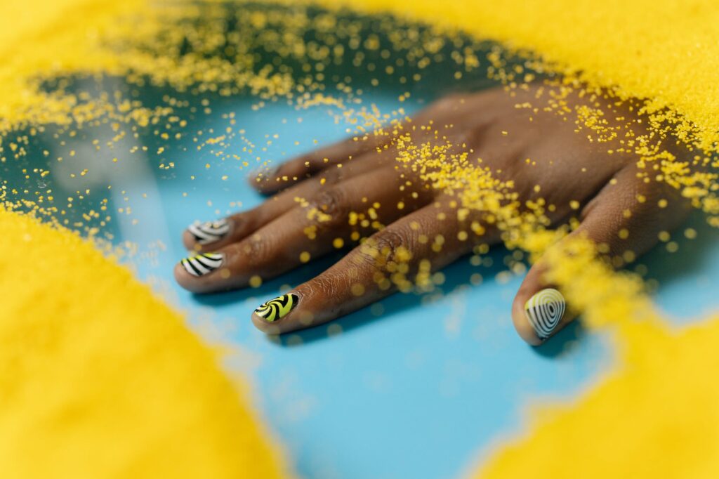 Uma mão com esmalte nas cores branca, preta e amarela sobreposta em uma superfície azul e alguns detalhes em amarelo. Imagem ilustrativa do texto sobre limpar mancha de esmalte do sofá.