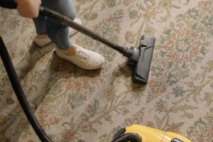 Pessoa limpando um carpete bege com o uso de um aspirador amarelo. Ilustração do texto sobre limpeza de carpete profissional.